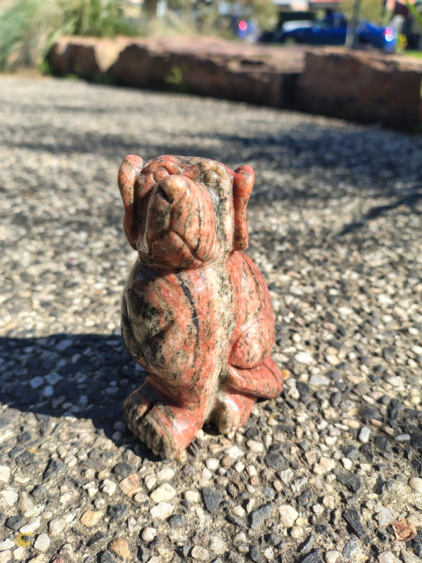 Unakite dog carving with natural green and pink tones, displayed on a rock outdoors.