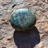 Labradorite palm stone with blue-green flashes and natural grey tones resting on a rock outdoors.