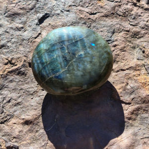 Labradorite palm stone with blue-green flashes and natural grey tones resting on a rock outdoors.